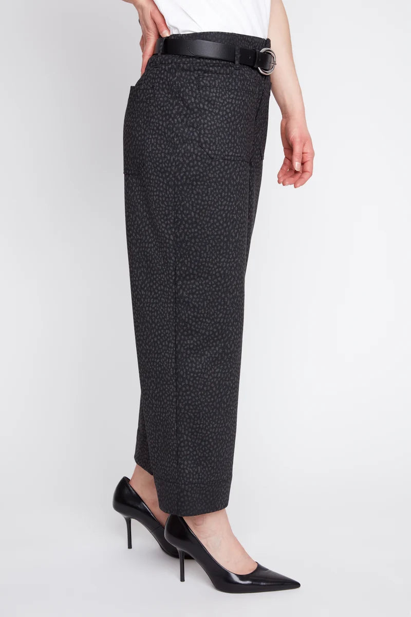 A model is shown from the waist down wearing black textured cropped trousers with a black belt, paired with black patent leather high heels against a light gray studio background.