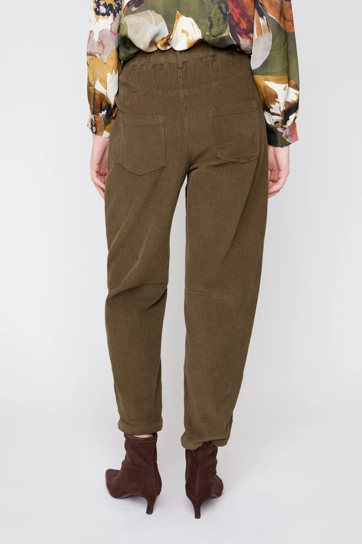 A person shown from the back wearing olive brown corduroy trousers, a multicolor floral long-sleeve top, and dark brown heeled ankle boots against a light gray studio background.