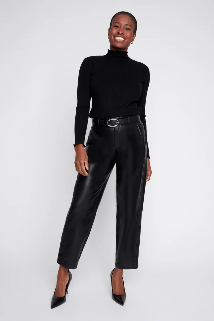 A model wearing a black long-sleeve mock neck top, black faux-leather cropped trousers with a black belt, and black pointed-toe heels, standing against a light gray studio background.