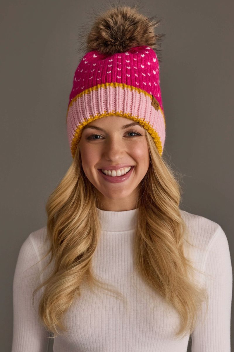 A smiling woman with long blonde hair wearing a pink and light pink knit beanie with a brown faux-fur pom-pom. The beanie features a white dotted pattern on the top section, a solid pink middle section, and a yellow-gold ribbed brim.