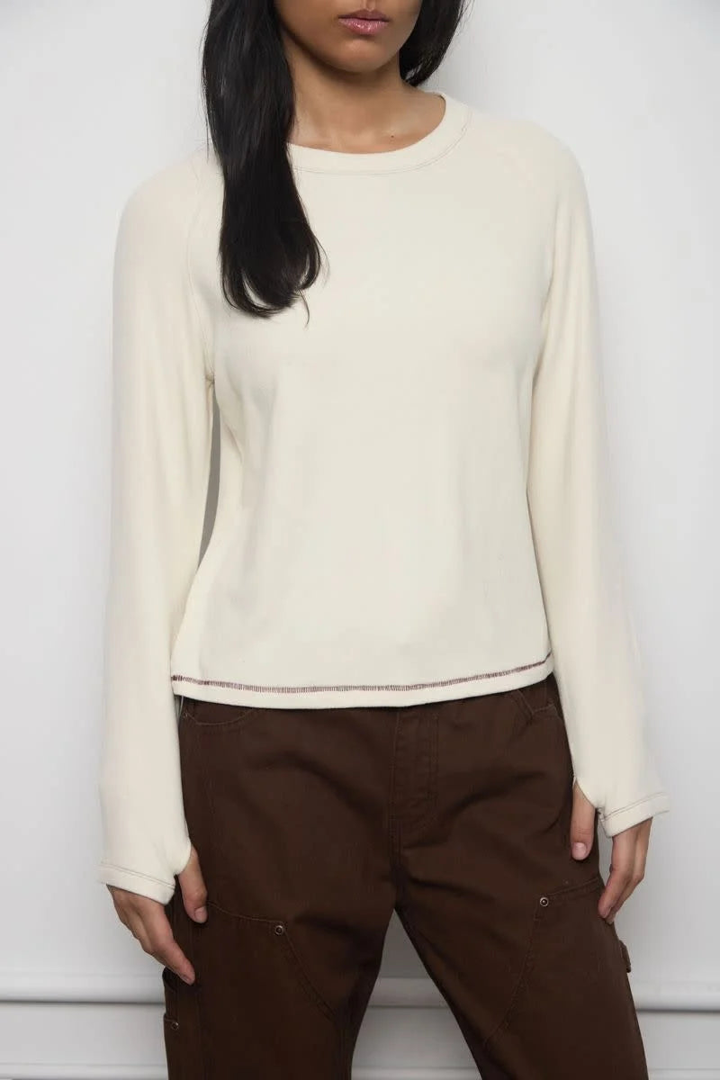 A woman wearing a cream-colored long-sleeve crewneck sweater and dark brown cargo pants, standing against a plain light gray wall.