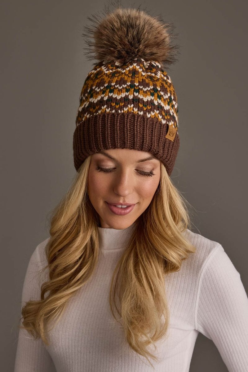 A woman wearing a brown knit beanie with a large brown faux-fur pom-pom and a multicolor patterned upper section, paired with a white ribbed turtleneck sweater against a solid gray background.