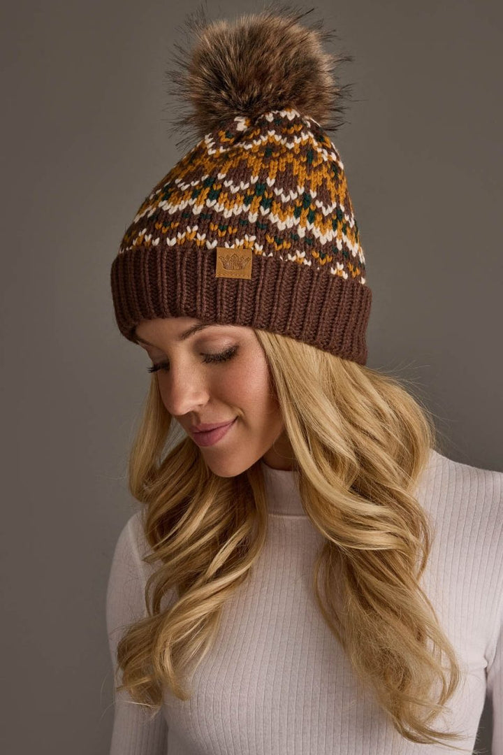 A woman wearing a brown knit beanie with a large brown faux fur pom-pom. The beanie features a multicolor Fair Isle pattern in mustard yellow, cream, and white above a solid dark brown ribbed brim.