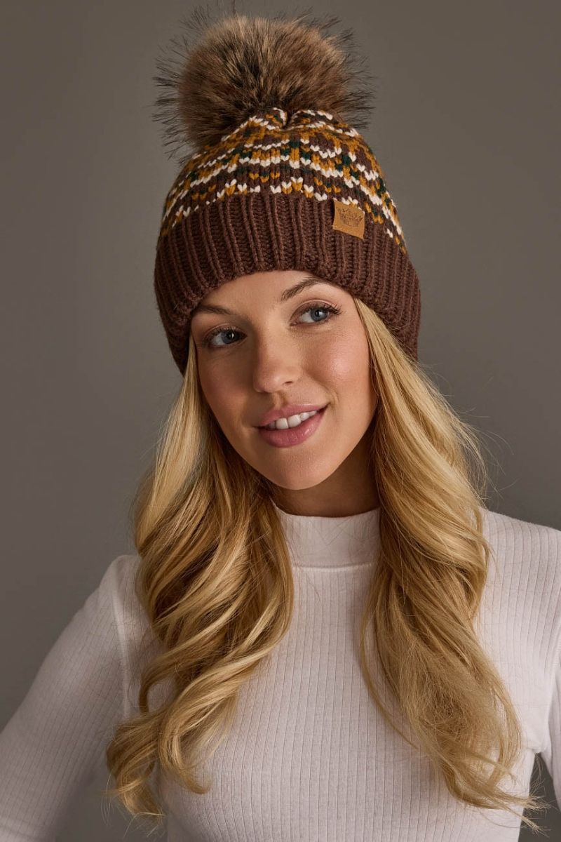 A woman wearing a brown knit beanie with a large brown faux-fur pom-pom and a multicolor knit pattern near the top. The beanie has a small tan leather patch on the side.