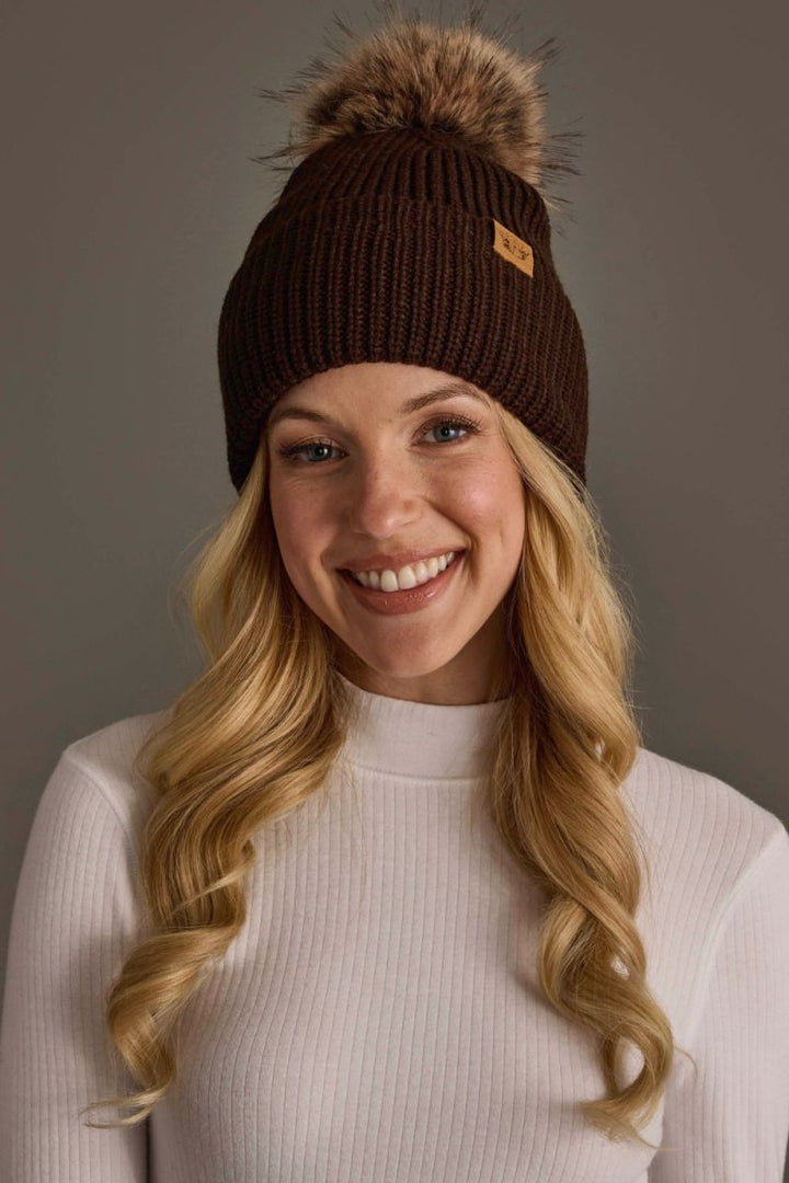 A smiling blonde woman wearing a dark brown ribbed knit beanie with a large brown faux-fur pom-pom and a small tan logo patch on the side, paired with a white ribbed mock-neck top against a solid gray studio background.