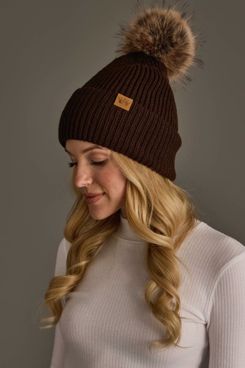 A woman wearing a dark brown ribbed knit beanie with a large brown faux-fur pom-pom and a small tan brand patch on the side, photographed against a plain gray studio background.