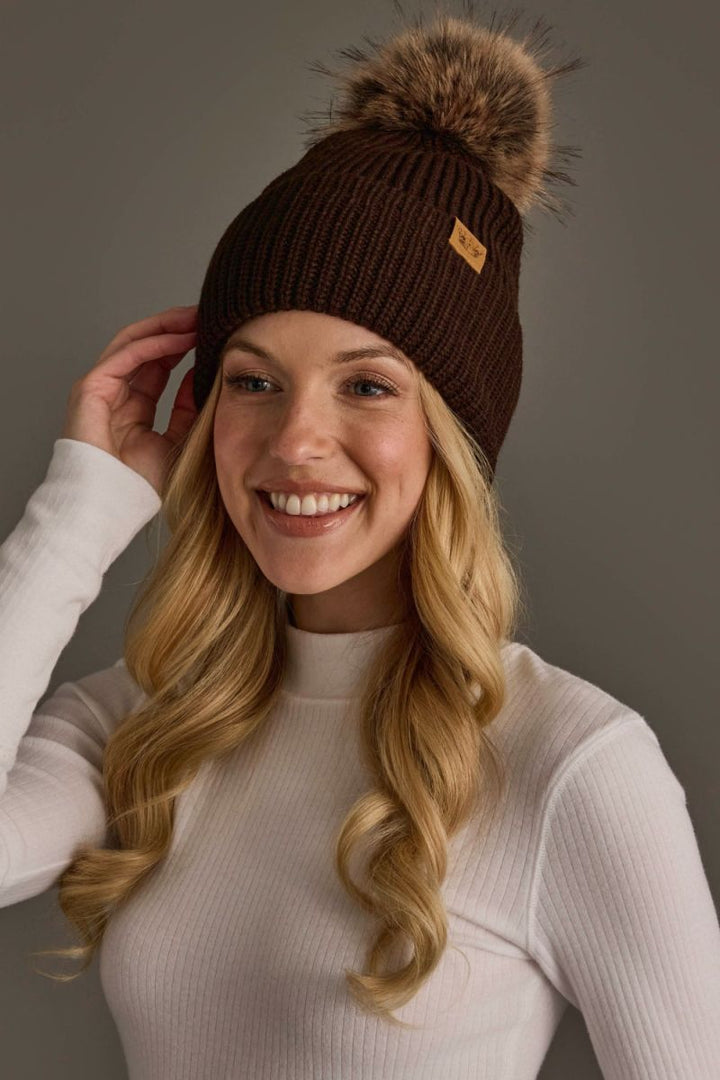 A smiling blonde woman wearing a dark brown ribbed knit beanie with a large brown faux-fur pom-pom and a small tan brand tab, photographed against a plain gray studio background.