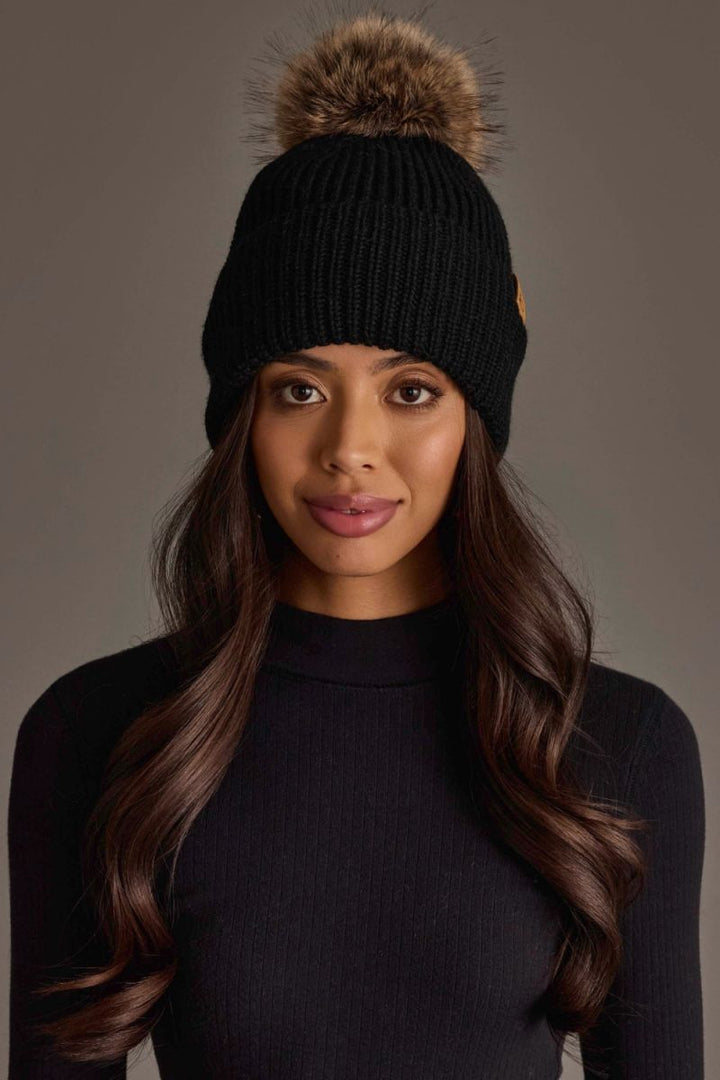 A woman wearing a black ribbed knit beanie with a brown faux-fur pom-pom and a small gold logo patch on the side, photographed against a plain taupe background.