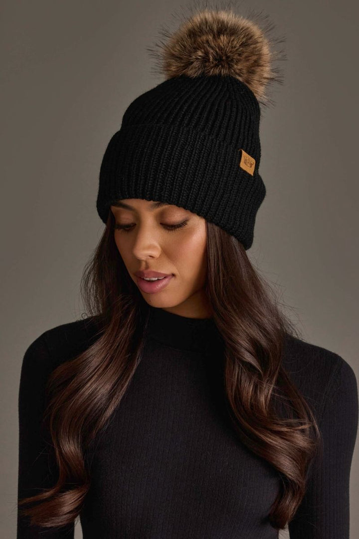 A woman wearing a black ribbed knit beanie with a large brown faux-fur pom-pom and a small tan brand patch on the side, photographed against a plain taupe background.
