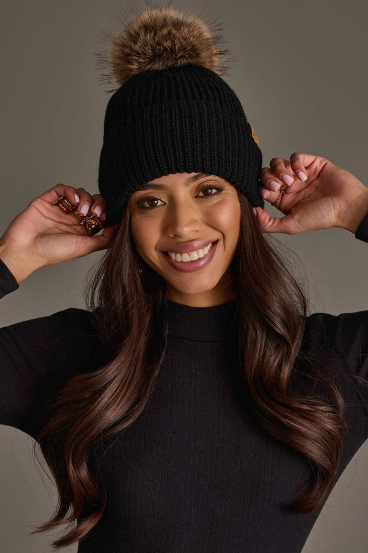 A smiling woman wearing a black ribbed knit beanie with a large brown faux-fur pom-pom and a small gold logo patch on the side, posed against a neutral gray studio background.