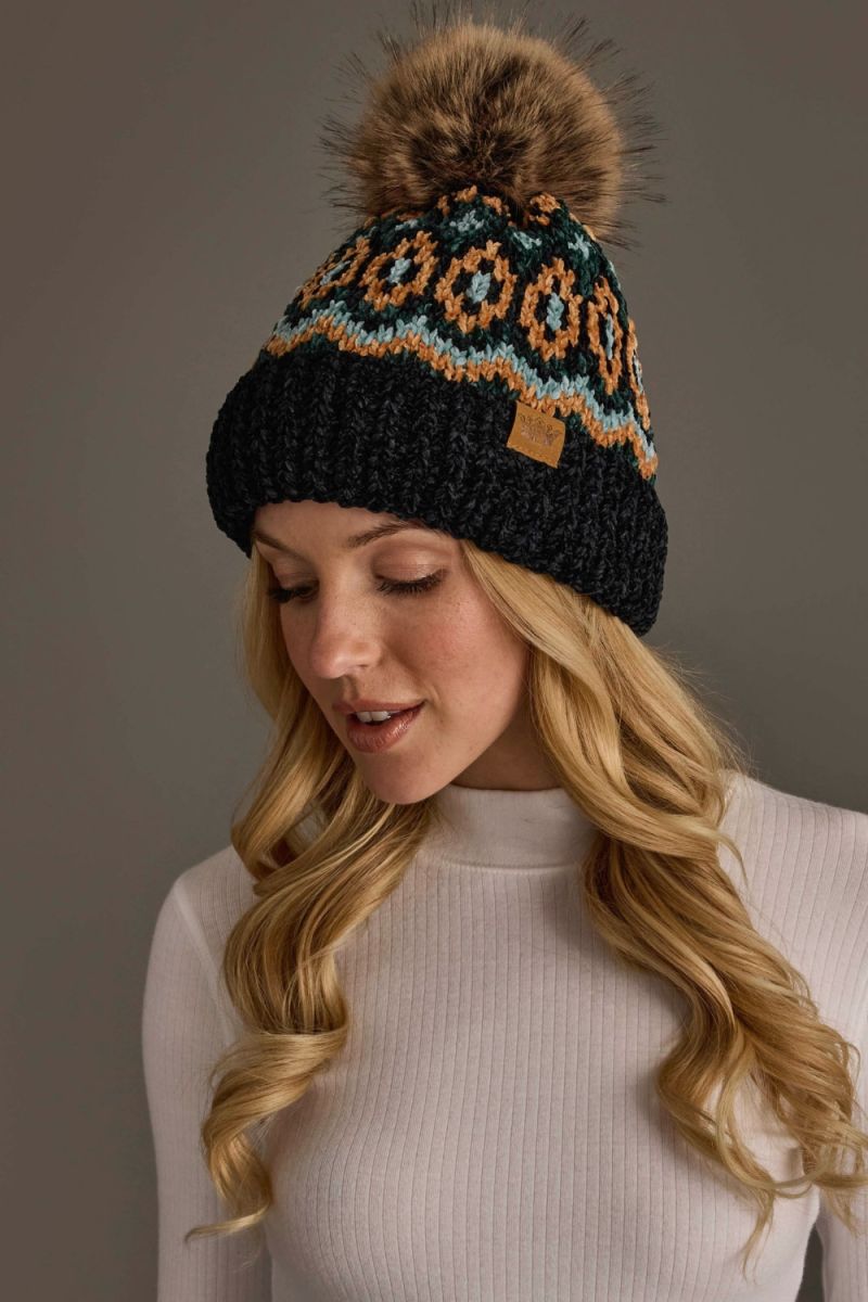 A woman wearing a black knit beanie with a brown faux-fur pom-pom and a colorful geometric pattern in orange, teal, and cream. The beanie has a small tan leather brand tag on the side.