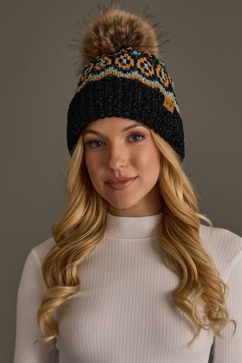 A woman with long blonde hair wearing a black knit beanie with a brown faux fur pom-pom and a colorful geometric pattern near the top, paired with a white ribbed mock neck top against a solid gray background.