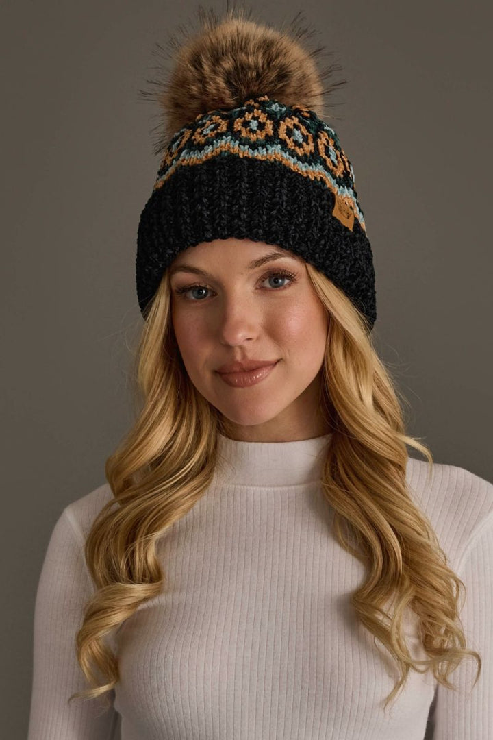 A woman with long blonde hair wearing a black knit beanie with a brown faux fur pom-pom and a colorful geometric pattern near the top, paired with a white ribbed mock neck top against a solid gray background.