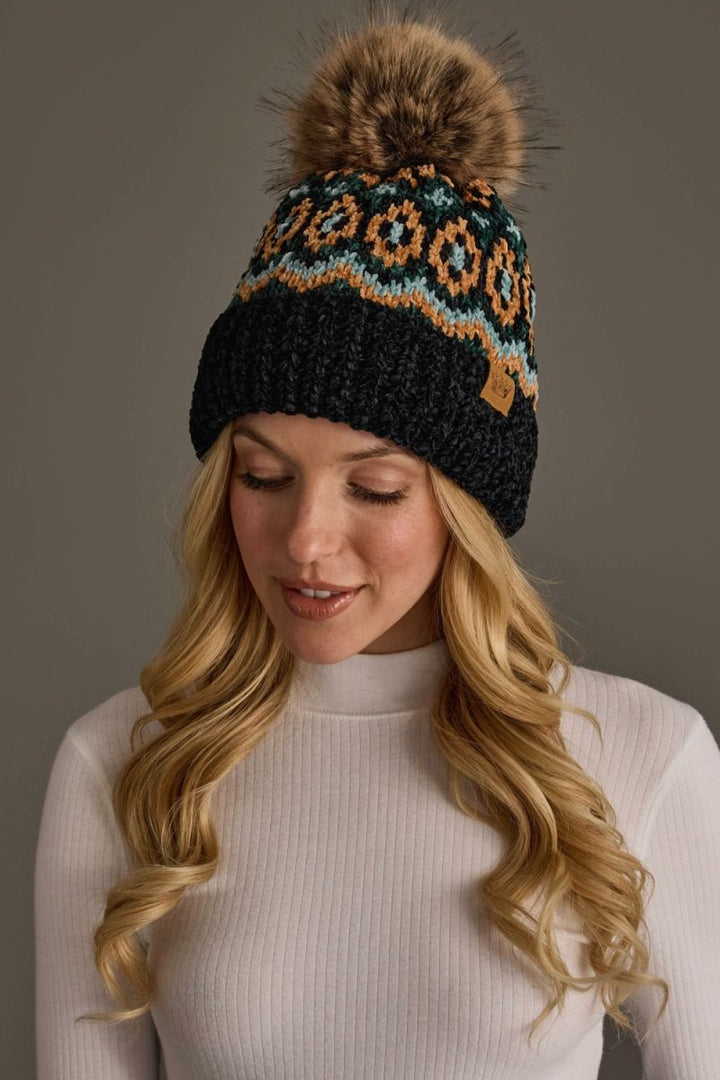 A woman wearing a black knit beanie with a brown faux-fur pom-pom and a colorful geometric pattern in orange, teal, and cream. She is wearing a white ribbed turtleneck sweater against a solid dark gray studio background.