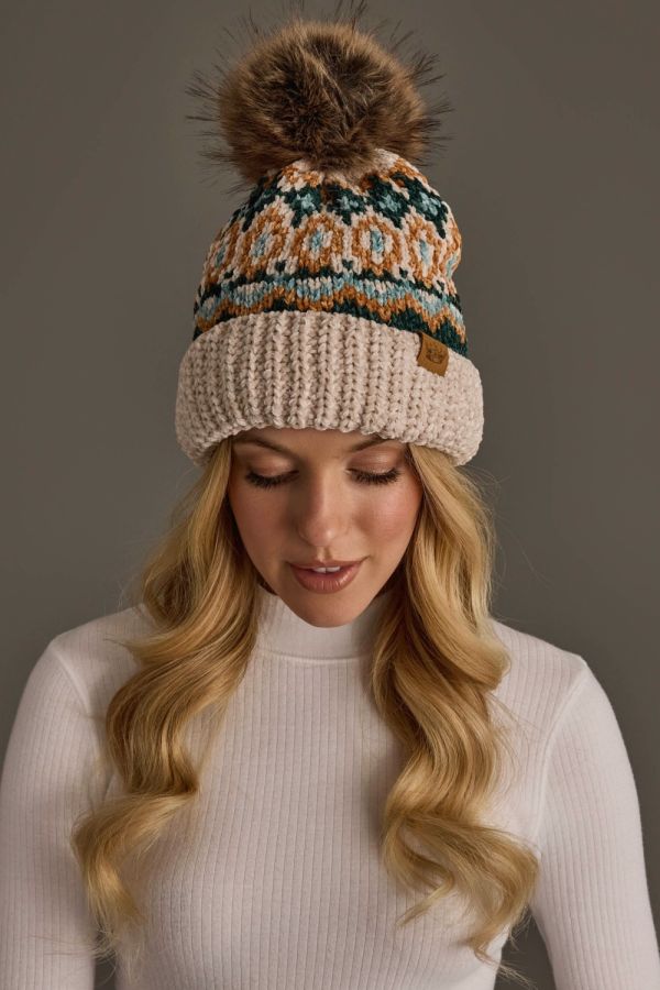 A woman wearing a cream knit pom-pom beanie with a multicolor Fair Isle pattern in teal, orange, and brown, paired with a white ribbed turtleneck sweater against a solid gray background.