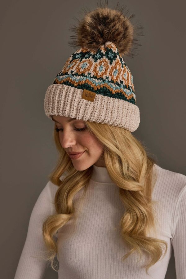 A woman wearing a cream knit beanie with a brown faux fur pom-pom and a multicolor geometric pattern on the crown. The beanie features a ribbed cuff and a small brown leather brand patch.