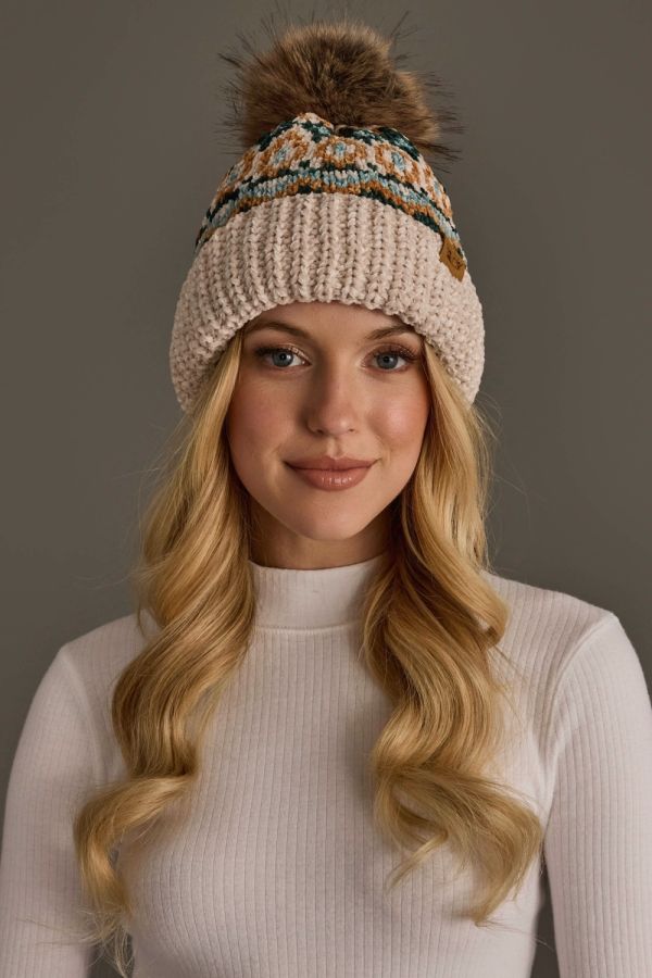 A woman with long blonde hair wearing a cream-colored knit beanie with a brown faux-fur pom-pom and a colorful patterned band, paired with a cream ribbed turtleneck sweater against a solid gray studio background.