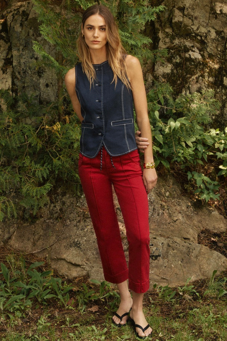 Woman in a blue sleeveless top and red pants standing against a stone wall with greenery.