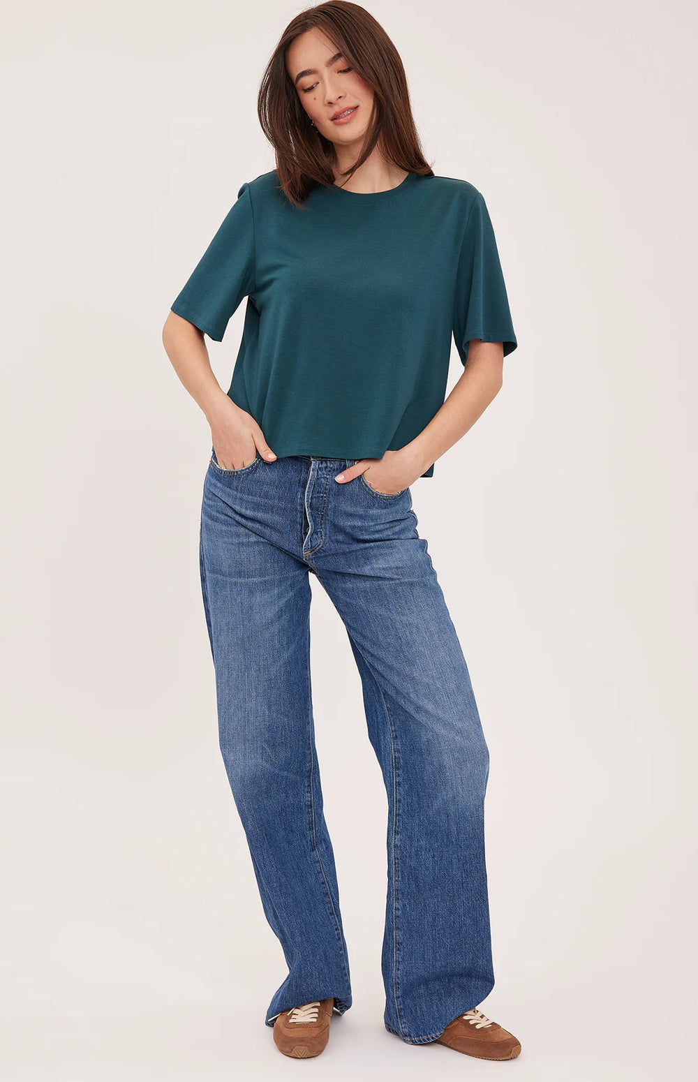 A woman modeling a dark teal green short-sleeve top and blue wide-leg jeans against a plain light background.