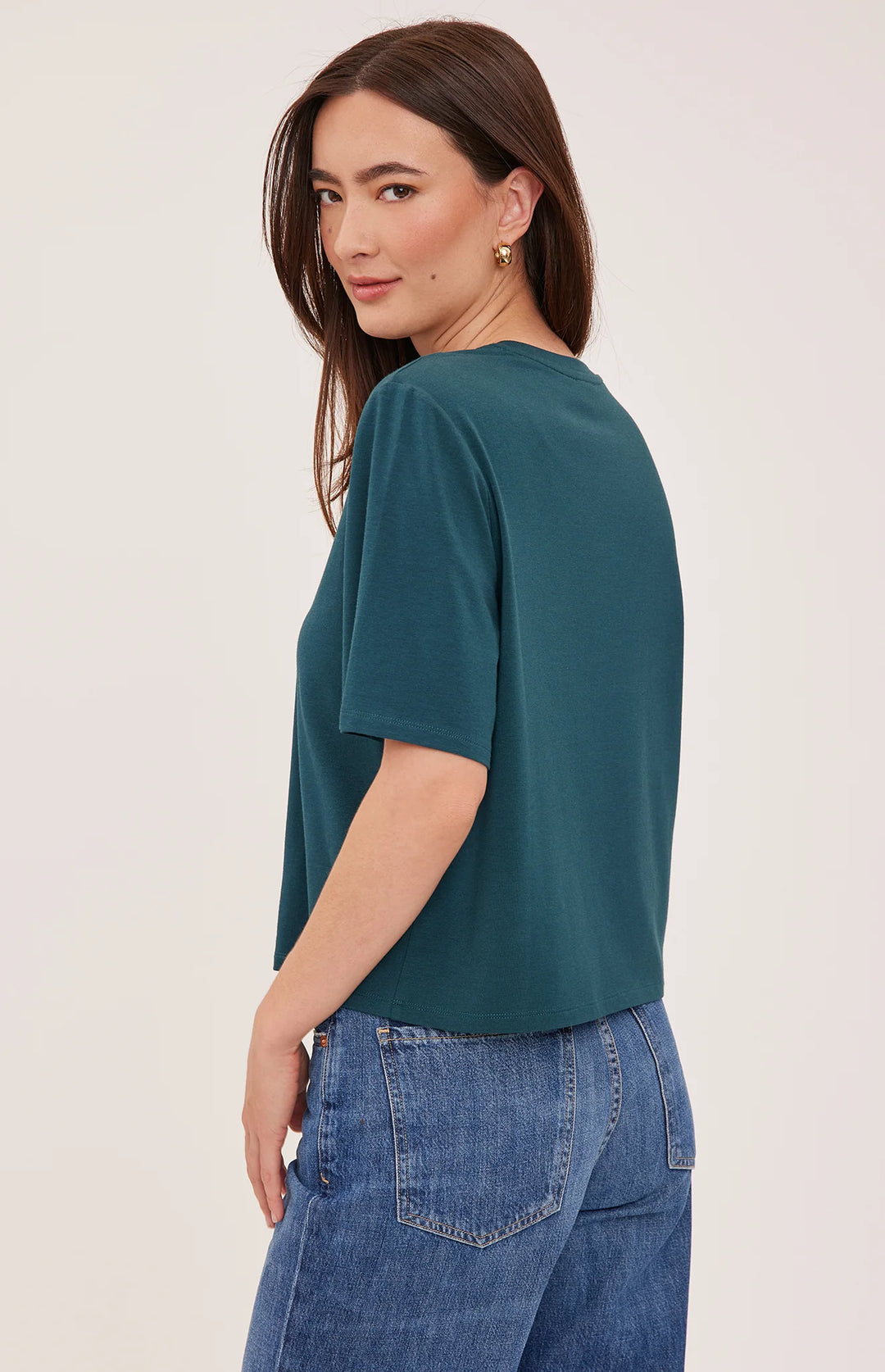 A woman wearing a short-sleeve crewneck top in teal green, paired with blue jeans, photographed from a side-back angle against a plain light background.