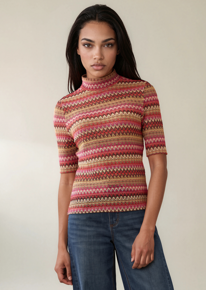 A woman modeling a short-sleeve mock-neck knit sweater with horizontal multicolor geometric stripes in pink, red, beige, and brown, paired with blue jeans against a plain beige studio background.