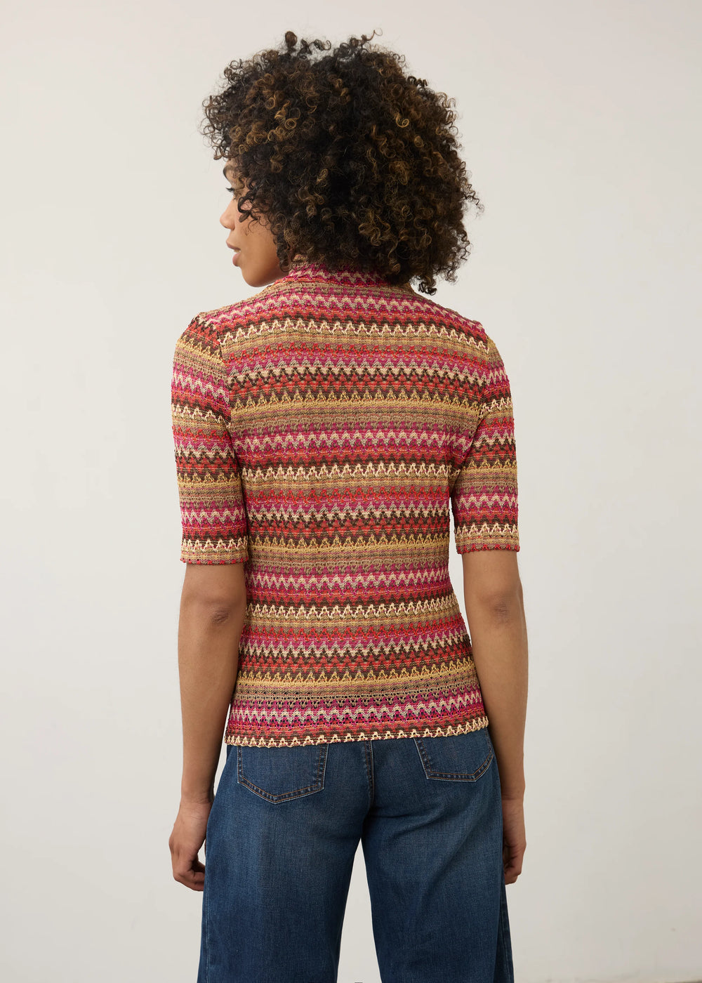A woman with curly dark hair is shown from the back wearing a short-sleeve knit top with horizontal multicolor geometric stripes in pink, red, beige, and brown, paired with blue jeans against a plain off-white background.