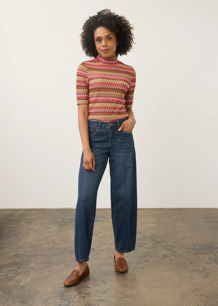 A woman stands in a studio wearing a short-sleeve mock-neck knit top with horizontal multicolor stripes in red, orange, yellow, green, and brown, paired with dark blue straight-leg jeans and brown leather loafers.