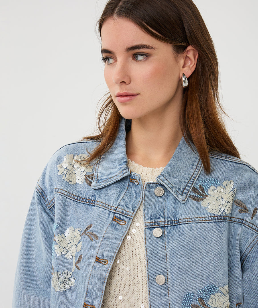 Woman wearing a denim jacket with floral embroidery on a plain background