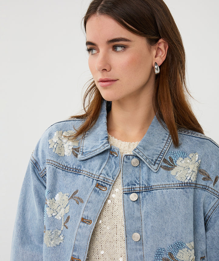 Woman wearing a denim jacket with floral embroidery on a plain background