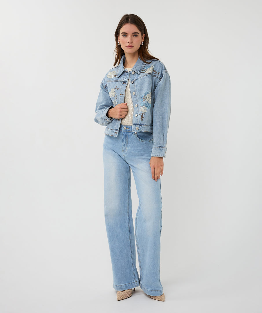 Woman wearing a light blue denim jacket with floral embroidery and matching jeans on a white background