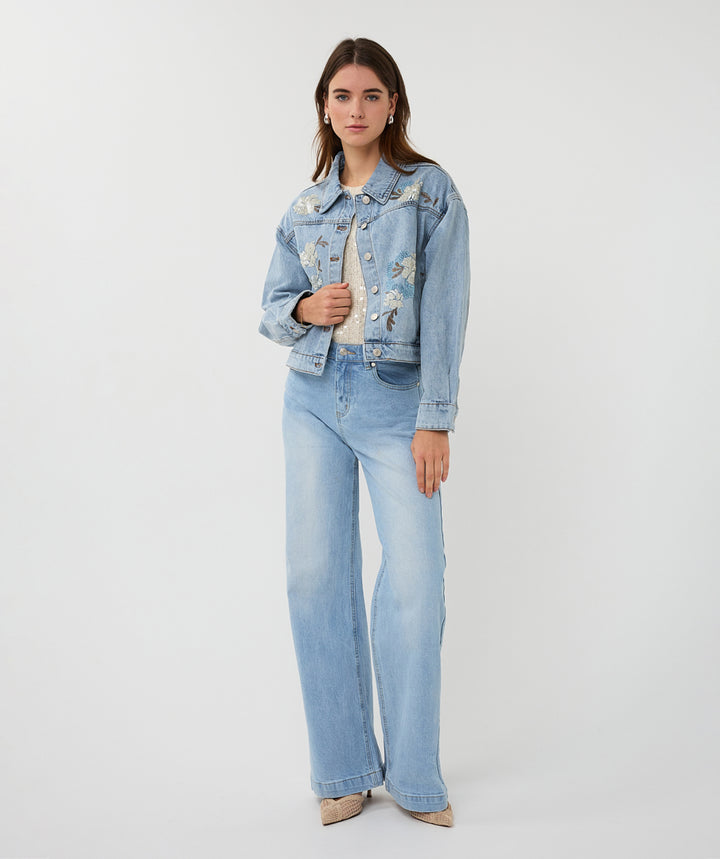 Woman wearing a light blue denim jacket with floral embroidery and matching jeans on a white background