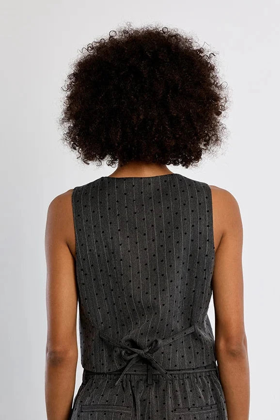 A woman viewed from behind wearing a dark gray sleeveless romper with a small repeating dot pattern and a tie detail at the back waist, against a plain light gray studio background.