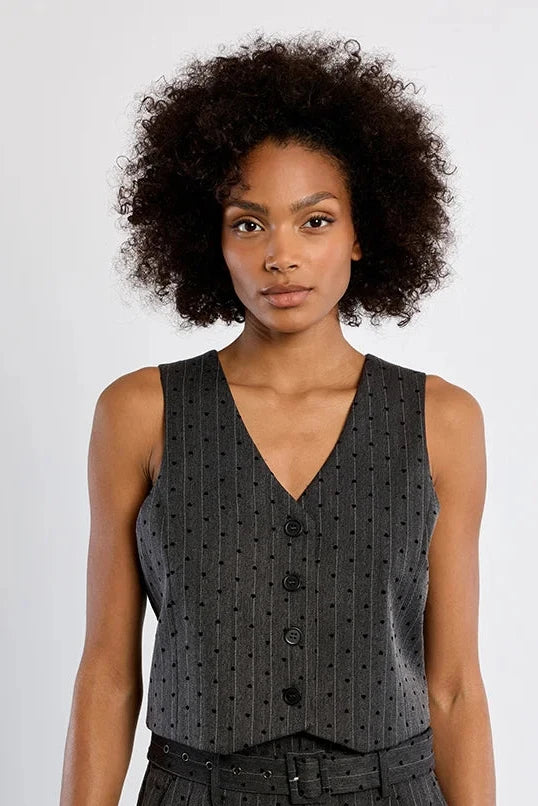 A model with curly dark hair wearing a dark gray sleeveless vest and matching shorts with a small white polka dot pattern, standing against a light gray studio background.