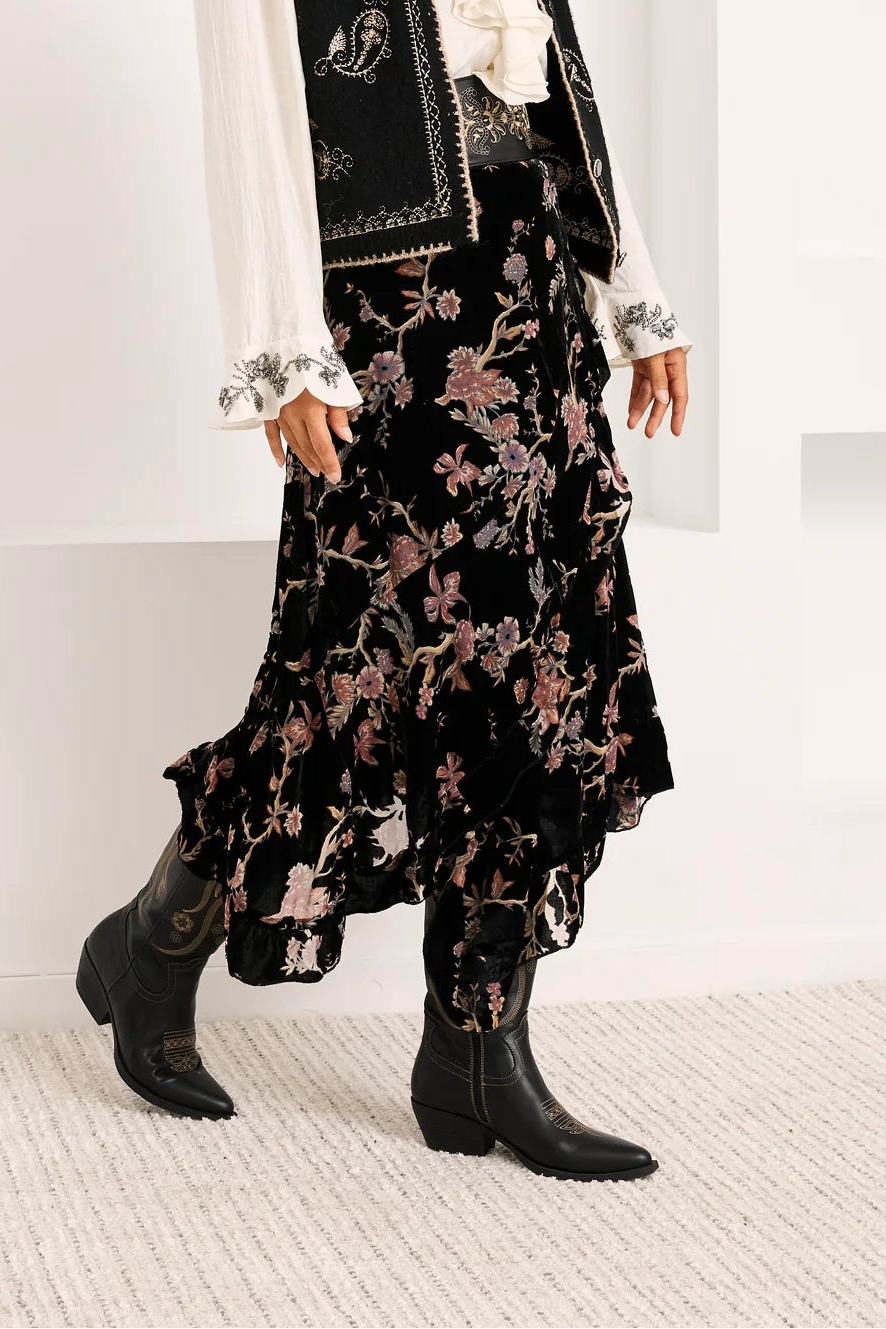 A woman models a bohemian-style outfit consisting of a black embroidered vest, a white ruffled blouse, and a black floral skirt, paired with black ankle boots, set against a white arched studio backdrop.