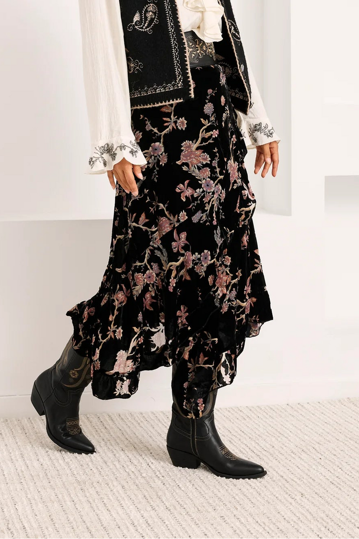 A woman models a bohemian-style outfit consisting of a black embroidered vest, a white ruffled blouse, and a black floral skirt, paired with black ankle boots, set against a white arched studio backdrop.