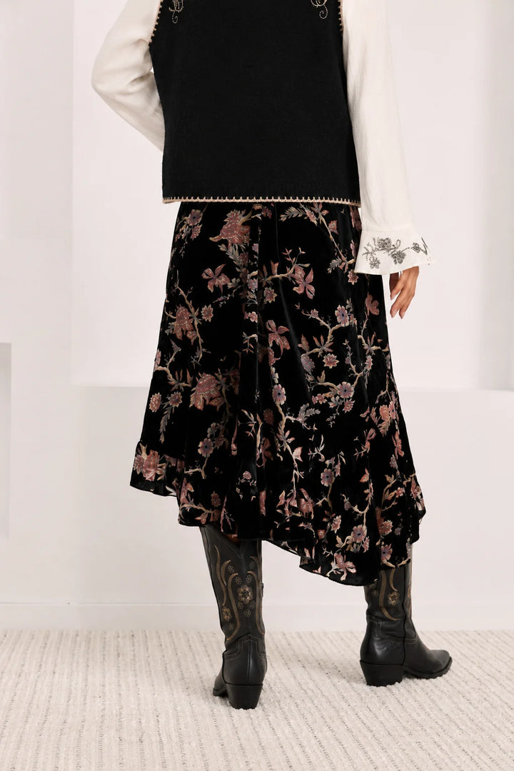 A person shown from the back wearing a black floral midi skirt with a black vest, white lace-sleeve blouse, and black cowboy boots in a bright studio setting.