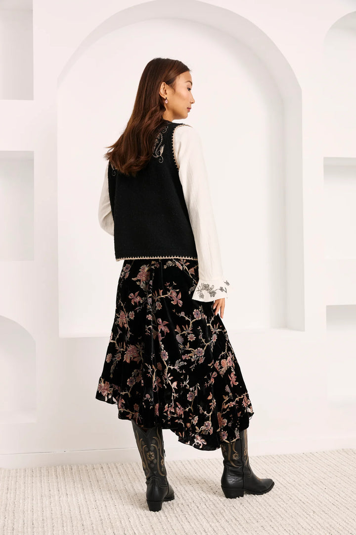 A woman is shown from behind wearing a black sleeveless vest over a white long-sleeve top, paired with a black velvet midi skirt featuring a pink floral pattern, and black cowboy boots, set against a white arched studio backdrop.