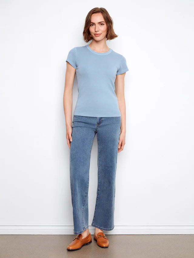 Woman wearing a light blue t-shirt and jeans standing against a white wall.