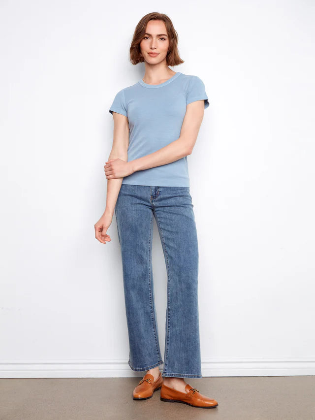 Woman wearing a light blue t-shirt and jeans standing against a white wall.