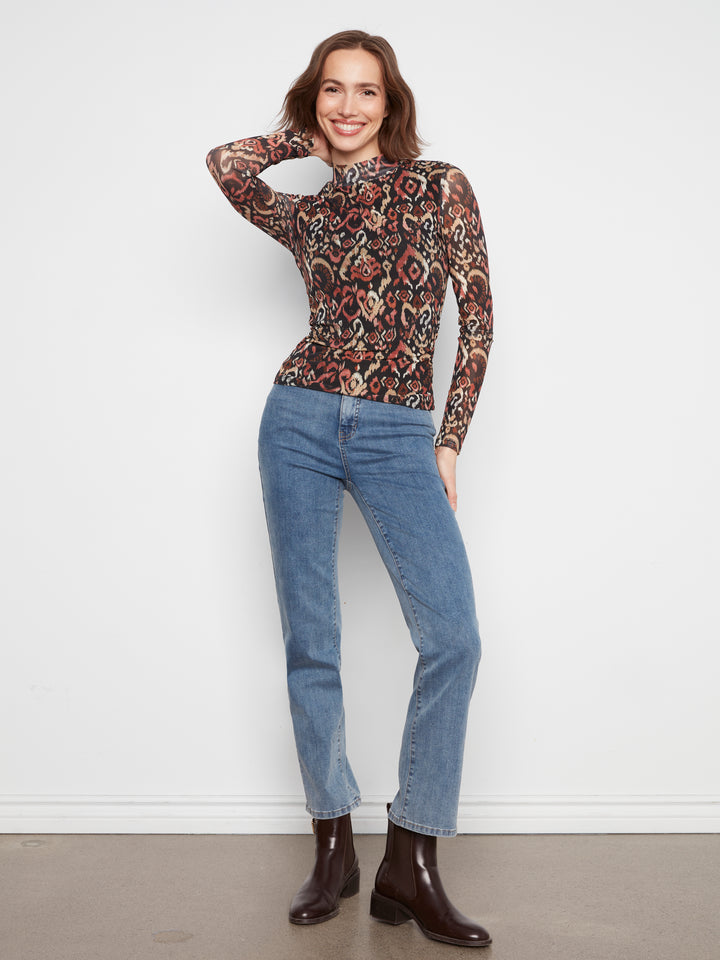 A woman modeling a long-sleeve mock neck top with a complex multicolor geometric and tribal pattern in red, brown, black, and beige, paired with blue jeans and dark brown ankle boots, standing against a white wall.