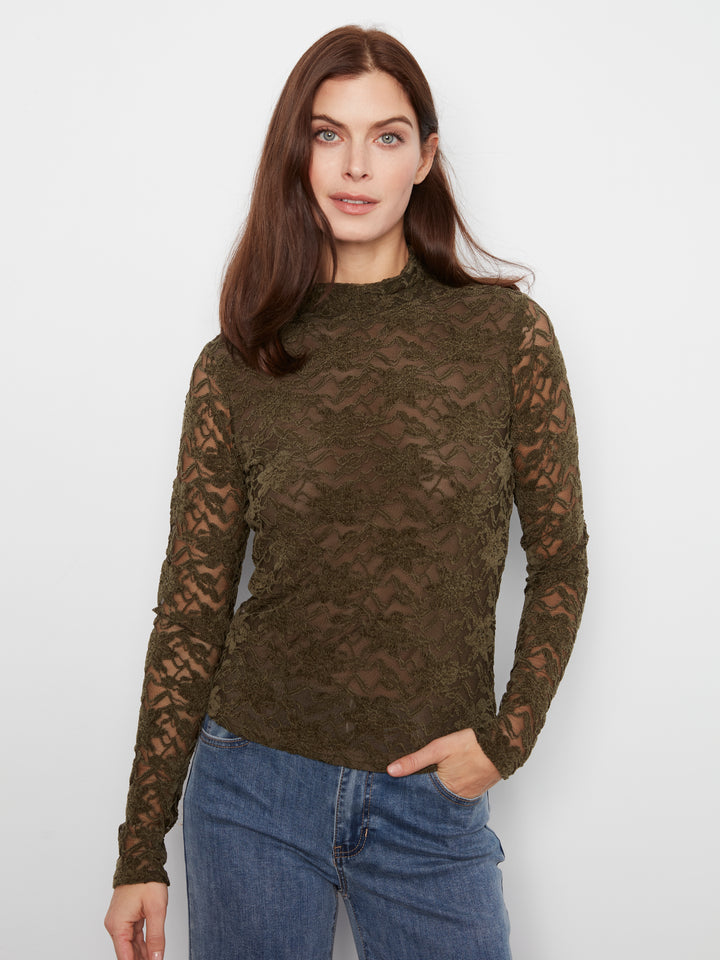 A woman wearing a dark olive green lace long-sleeve mock neck top and blue jeans, standing against a plain light gray background.