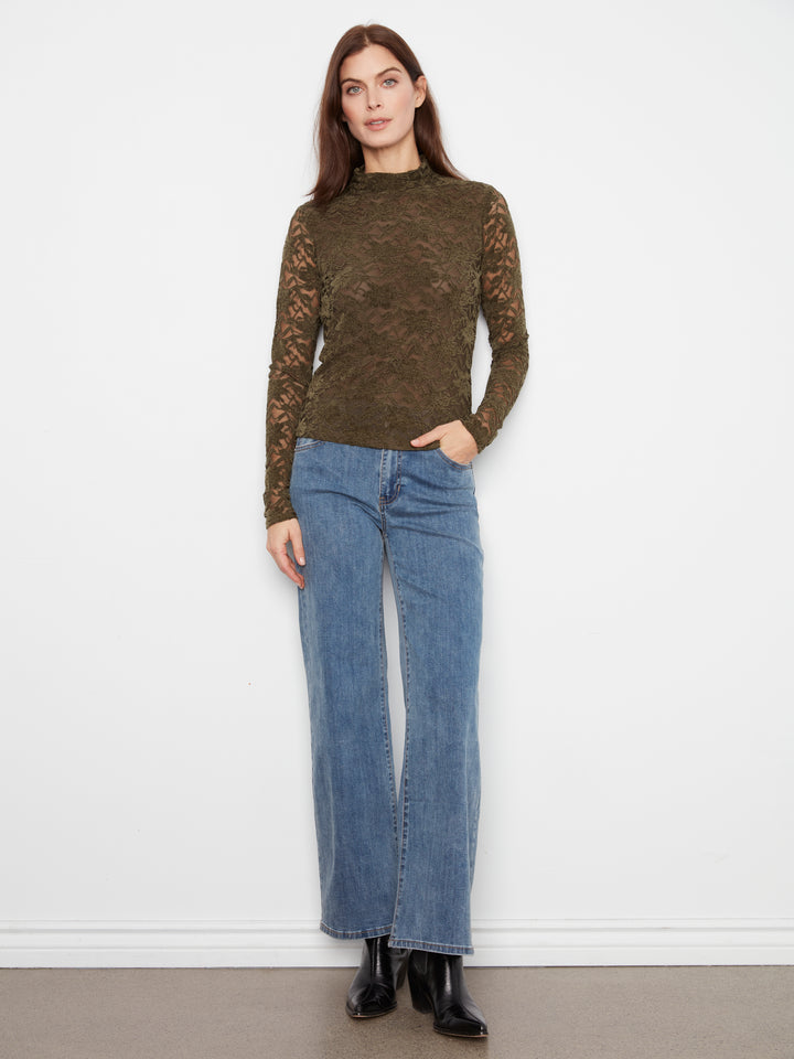 A woman modeling a dark olive green lace long-sleeve top with blue wide-leg jeans and black ankle boots, standing against a plain white wall.
