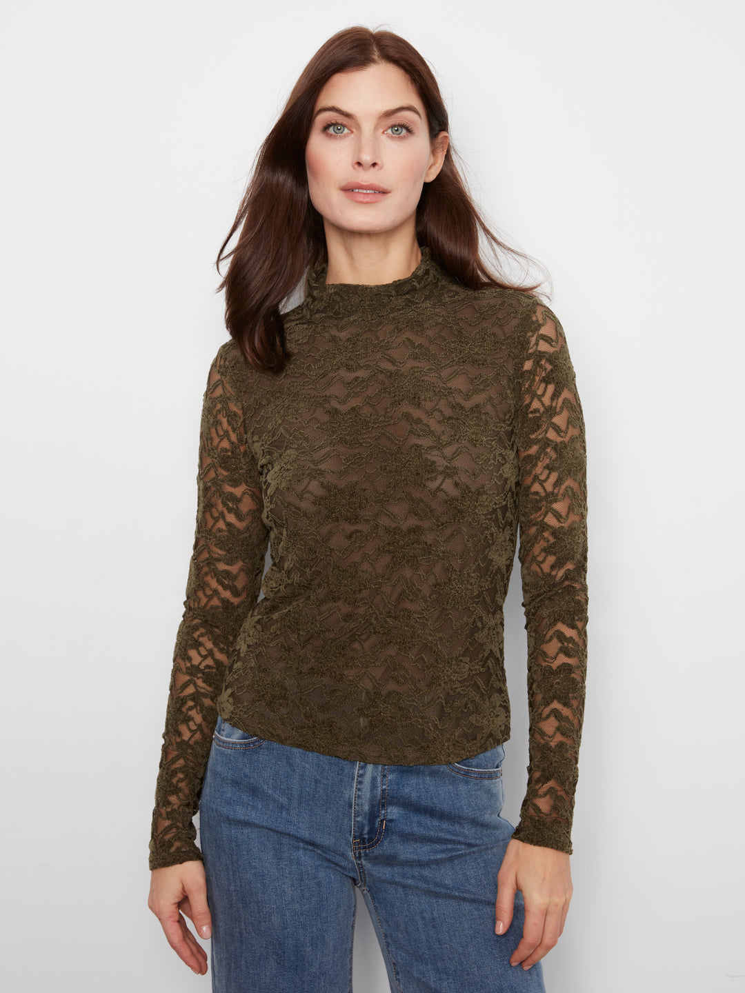 A woman wearing a dark olive green lace long-sleeve mock neck top and blue jeans, standing against a plain white studio background.