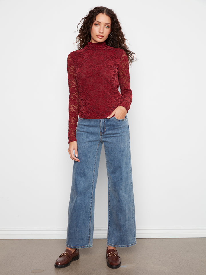A woman modeling a long-sleeve burgundy lace mock neck top paired with wide-leg blue jeans and brown leather loafers, standing against a plain white wall.