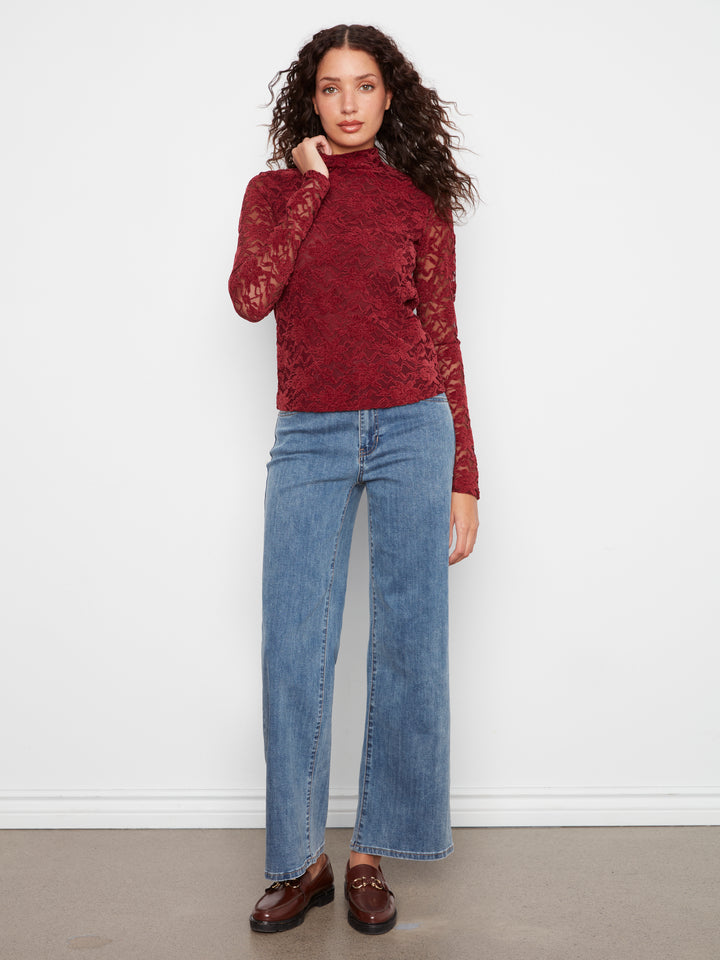 A woman modeling a long-sleeve burgundy lace mock-neck top paired with wide-leg blue jeans and brown leather loafers, standing against a white wall.