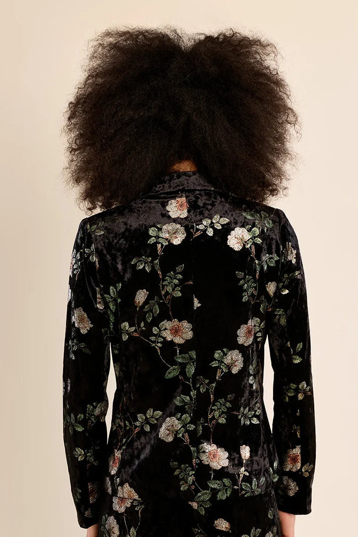 A person shown from the back wearing a black velvet long-sleeve blazer with an embroidered floral pattern in green, pink, and white, set against a solid beige background.