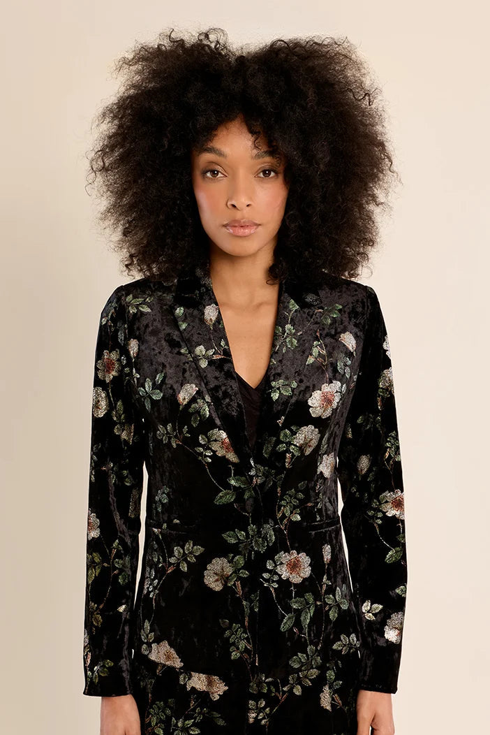 A model with voluminous dark curly hair wearing a black velvet long-sleeve wrap dress with a muted floral pattern, set against a plain beige studio background.