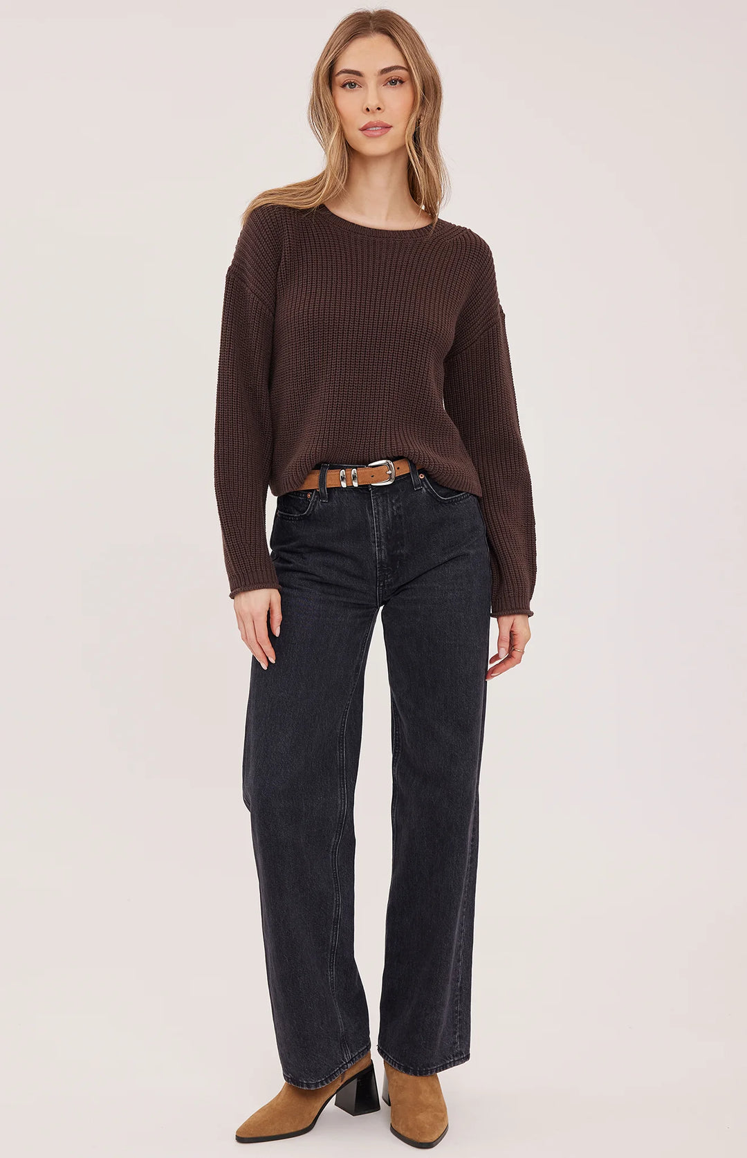 A woman modeling a dark brown ribbed knit sweater, dark wash wide-leg jeans, a brown leather belt, and brown suede boots against a plain light background.