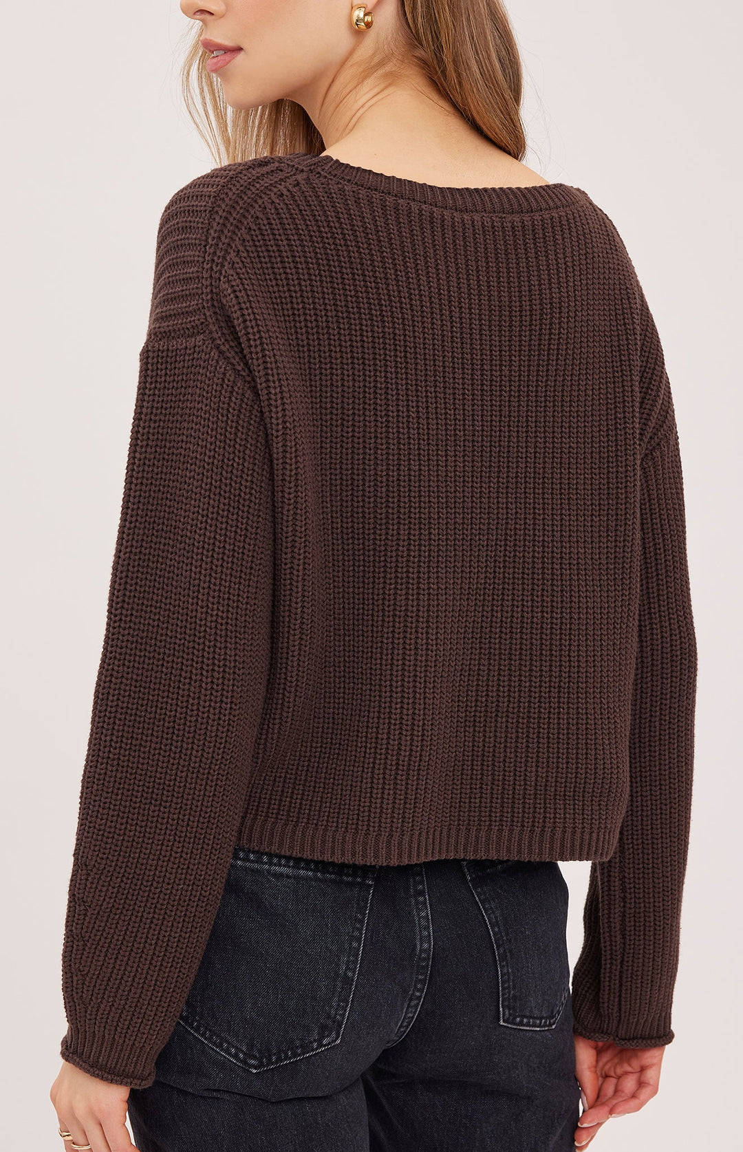 A woman wearing a dark brown ribbed knit sweater, shown from the back. The sweater has a wide boat neckline and long sleeves, paired with dark wash jeans against a plain light background.