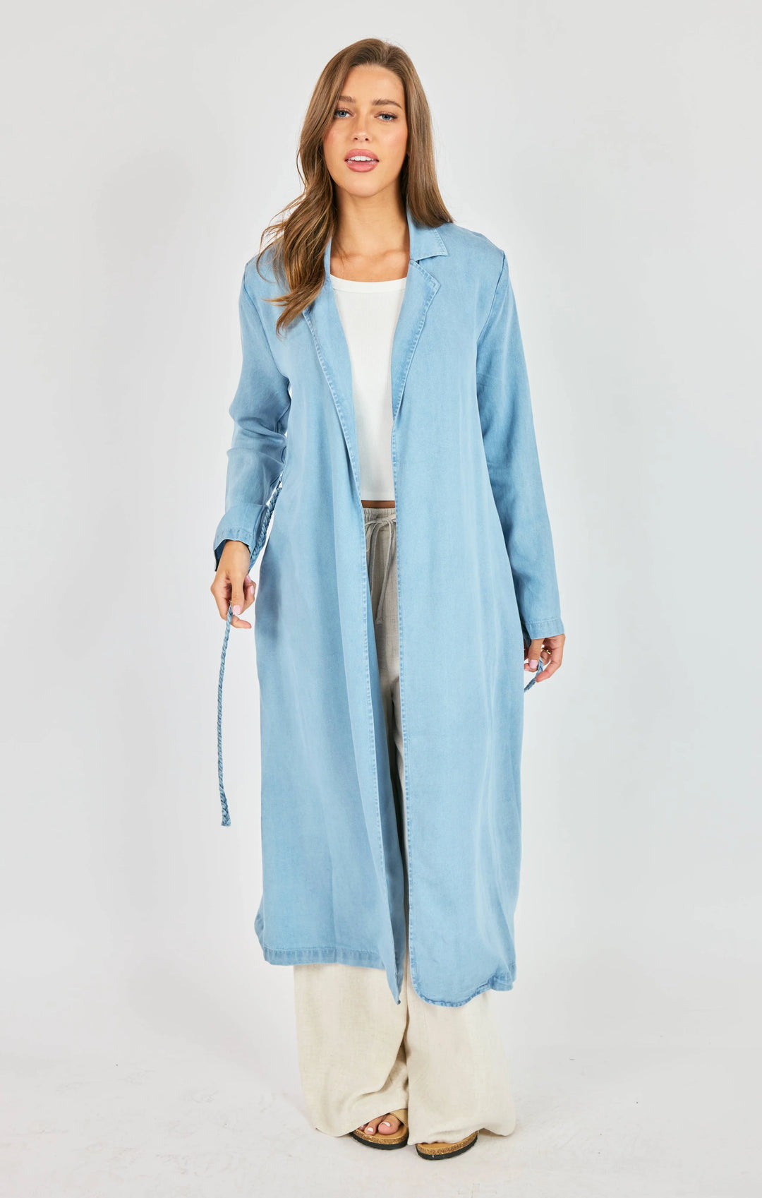 Woman wearing a long light blue cardigan over a white top and pants on a gray background